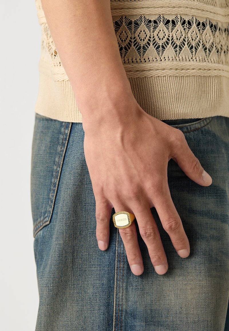 Gylden metallring med et rektangulært, merket topp og teksturerte sider, båret på en hånd, kombinert med en strikket beige topp og denim jeans.