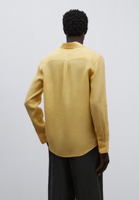 Camisa amarilla de manga larga con yugo trasero, dobladillo redondeado y puños abotonados. Confeccionada con una tela ligera y un acabado ligeramente texturizado.