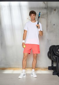 Sportaš u bijeloj majici s kratkim rukavima i koraljnim šortsevima, drži tenisku reketa i lopticu, s bijelim tenisicama i ručnim trakama. Teretana.