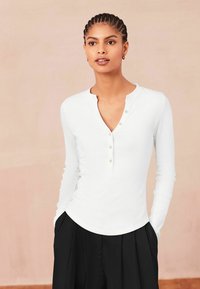 Bijela dugorukava henley majica s V-izrezom, tri gumba i rebrastom teksturom, u kombinaciji s crnim naboranim hlačama.