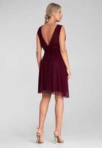 Burgunderfarbenes Kleid mit tiefer V-Ausschnitt am Rücken, gerafftem Taillenbereich und fließendem Rock. Leichter Stoff mit transparenter Überlagerung. Getragen mit silbernen High Heels.