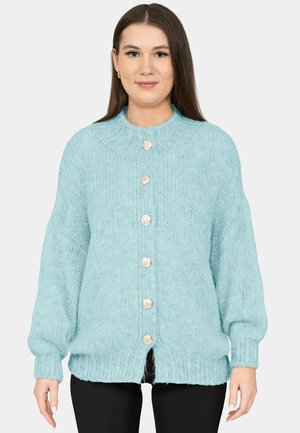 Vrouw draagt een lichtblauwe grofgebreide cardigan met zes gouden knopen, gecombineerd met zwarte broek, staand tegen een effen achtergrond.