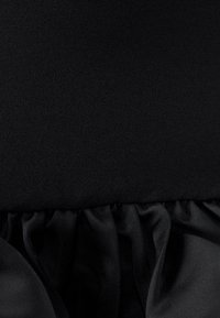 Black fabric garment with a smooth, textured surface, featuring gathered ruffles along the bottom edge. Simple, monochromatic design.