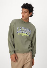 Sudadera de color verde oliva con puños y dobladillo acanalados, que presenta un gran logo "LACOSTE" y un gráfico de cocodrilo en colores contrastantes.