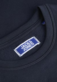 Granatowa bluza crewneck z prążkowanym dekoltem. Posiada prostokątną metkę marki w niebiesko-białych kolorach, wszytą w wewnętrzny kołnierz.