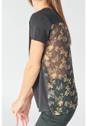 Kvinde iført en sort t-shirt med korte ærmer og et gennemskinneligt panel med blomsterprint på ryggen, kombineret med mørke bukser.