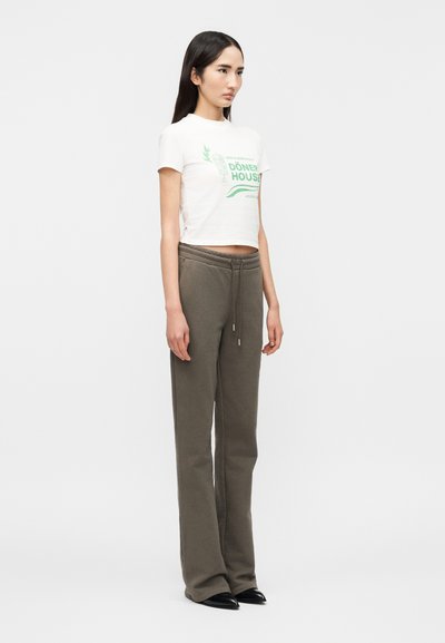 Νεαρή γυναίκα όρθια, με λευκό κροπ τοπ με  πράσινο κείμενο και χακί παντελόνι φόρμας με ρίγα, φορώντας μαύρα μυτερά παπούτσια.