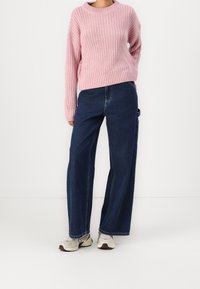 Jasnoróżowy, dziergany sweter z okrągłym dekoltem, w zestawieniu z jasnoniebieskimi, szerokimi jeansami z wysokim stanem oraz beżowymi sportowymi butami z ciemnymi akcentami.