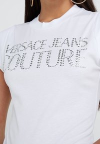 Bílá bavlněná tričko s krátkým rukávem a klasickým kulatým výstřihem, na kterém je nápis "VERSACE JEANS COUTURE" vyrobený ze stříbrných rhinestone kamínků.