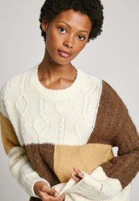 Adīts džemperis ar krāsu blokiem krēmkrāsā, brūnā un bēšā. Iezīmes kabelīšu adījuma raksti un ribotas aproces; mīksta, teksturēta auduma.