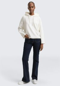 Weiße Kapuzenjacke mit geripptem Muster, kombiniert mit dunkelblauen Schlagjeans und weißen Turnschuhen. Das Modell steht vor einem neutralen Hintergrund.