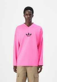 Långärmad rosa sporttröja med strukturerad tyg, med en svart Adidas-logotyp i mitten och V-ringning.