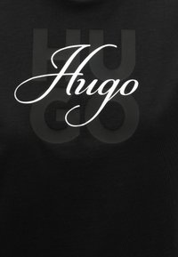 Μαύρο ύφασμα με μεγάλο σκούρο γκρι κείμενο "HUGO" στο φόντο και λευκό κείμενο με καλλιγραφική γραφή "Hugo" στο κέντρο.