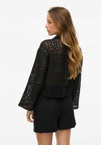 Crna čipkana jakna s dugim rukavima i cvjetnim uzorcima, koja ima otvoreni dizajn. U kombinaciji s odgovarajućim crnim šortsevima.