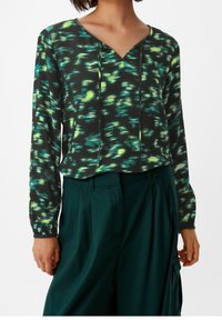 Schwarze Bluse mit grünem und gelbem abstraktem Muster, V-Ausschnitt, lange Ärmel mit elastischen Bündchen, kombiniert mit hoch taillierten grünen Hosen.