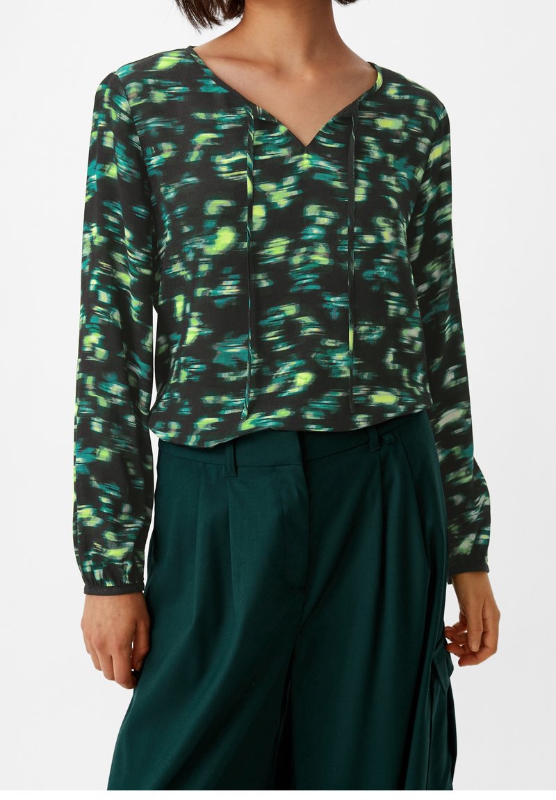 Schwarze Bluse mit grünem und gelbem abstraktem Muster, V-Ausschnitt, lange Ärmel mit elastischen Bündchen, kombiniert mit hoch taillierten grünen Hosen.