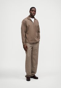 Καφέ πουλόβερ με φερμουάρ, ελαφρύ ύφασμα, σε συνδυασμό με ανοιχτό καφέ παντελόνι με υφή. Σκούρα παπούτσια ολοκληρώνουν την εμφάνιση.