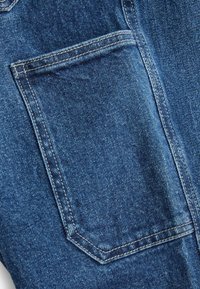 Sinine denim kangas, millel on tagatasku kolmnurga kujundusega, tugevdusõmblused ja tekstureeritud viimistlus.
