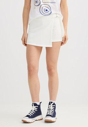 Mujer con una falda envolvente blanca texturizada con hebilla lateral, combinada con una camiseta gráfica blanca y zapatillas altas azul marino con cordones blancos.