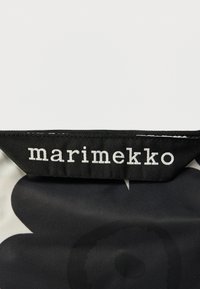 Sort-hvid Marimekko taske med blomstermønster. Materialet er glat stof med en robust sort lynlås og en fremtrædende mærkelap.