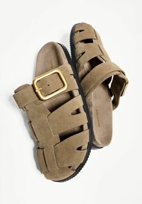 Rjave semišne natikače z širokim trakom, opremljene z zlatim sponko in odprto zasnovo. Teksturiran podplat z črnim podplatom.