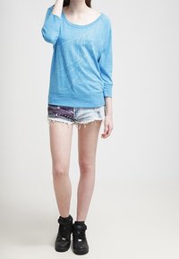 Ljusblå, långärmad sweatshirt med en stor vit Nike-logotyp. Kombination med slitna denimshorts och svarta högkanta sneakers.