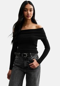 Schwarzes, schulterfreies Ripp-Oberteil mit langen Ärmeln, kombiniert mit hochgeschnittenen, dunklen Jeans und einem schwarzen Gürtel mit einer runden silbernen Schnalle.
