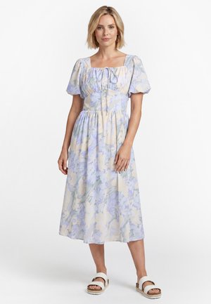 Kvinde iført en pastelblomstret midi-kjole med pufærmer og firkantet udskæring, kombineret med hvide slip-on sandaler, stående på hvid baggrund.