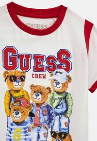 T-shirt bianca con bordini rossi che presenta una grafica cartone di orsi in abiti colorati, con la scritta "GUESS CREW" e accessori giocosi come occhiali da sole e cappelli.