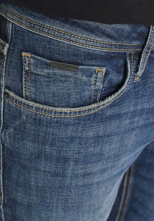 Blaue Jeans aus Denim mit einer Nahaufnahme von einer Tasche, die kontrastierendes Näht zeigt und ein schwarzes Etikett hat. Der Stoff hat eine strukturierte Oberfläche.