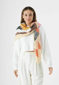 Écharpe à motifs géométriques multicolores en tissu doux, présentant des nuances de jaune, rose, marron et noir. Drapée élégamment sur un haut blanc.
