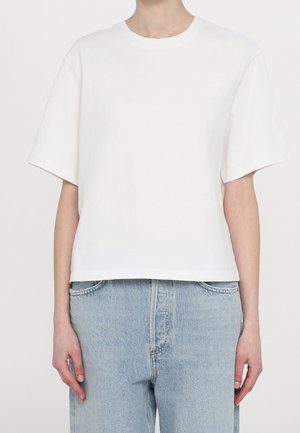 Person trägt ein schlichtes weißes T-Shirt mit kurzen Ärmeln und hellblaue hoch taillierte Jeans, steht vor einem schlichten Hintergrund.