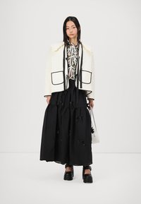 Λευκό τζάκετ με πλισέ σχέδιο και μαύρες λεπτομέρειες, συνδυασμένο με μια floral κορυφή με κορδόνια και μαύρη φούστα με φιόγκους. Χοντρές μαύρες παπουτσες ολοκληρώνουν το σύνολο.