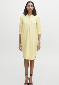 Robe en lin jaune avec un col mandarin, des manches trois quarts et des boutons sur le devant. Présente des fentes latérales à l'ourlet.