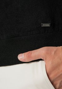Czarny sweter dziergany z ściągaczami, z małą srebrną metką. Dłoń spoczywa na białej kieszeni pod brzegiem swetra.