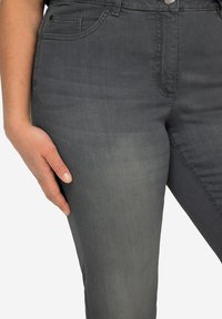 Jeans gris foncé avec une texture lisse, présentant un décoloration subtile. Des poches avant et un détail de fermeture à bouton sont visibles sur la taille.
