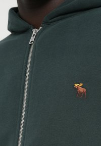 Haut de sweatshirt zippé de couleur vert foncé. Il est orné d'un petit orignal brodé en marron et jaune sur la partie gauche de la poitrine.