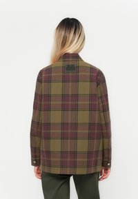 Person med långt, ljus ombre-hår bär en grön och lila rutig jacka med texten "CPH Studios" på övre delen av ryggen, tillsammans med mörka byxor.