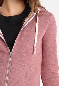 Persona vistiendo una sudadera con cremallera rosa sobre una camiseta negra de cuello redondo, con los cordones de la sudadera blanca visibles.