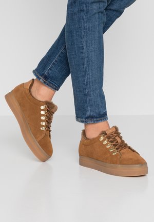 Braune Wildledersneaker mit Fellfutter und goldenen Ösen, getragen zu blauen Skinny-Jeans auf weißem Boden.