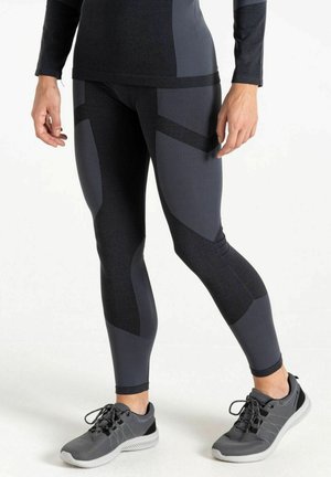 Persona que lleva leggings negros y gris oscuro con estampado y zapatos deportivos grises, de pie sobre una superficie blanca.