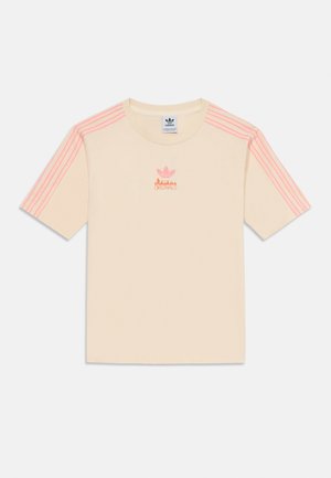 Kermanvärinen puuvillapaita, jossa on lyhyet hihat, ja siinä on vaaleanpunaiset kolmiraidalliset yksityiskohdat sekä brodeerattu vaaleanpunainen Adidas-logo rinnassa.