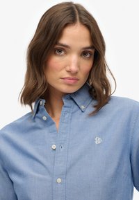 Jasnoniebieska koszula z kołnierzykiem, z subtelnymi detalami w szyciu, logo po lewej stronie klatki piersiowej oraz beżowymi guzikami.