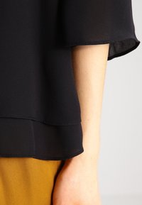 Blouse noire transparente à manches larges, design superposé à l'ourlet. Portée sur un vêtement couleur moutarde. Texture lisse et matériau léger.