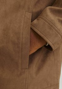 Chaqueta de ante marrón con un bolsillo lateral y puños de manga doblados, que muestra una textura suave y detalles de costura limpios.