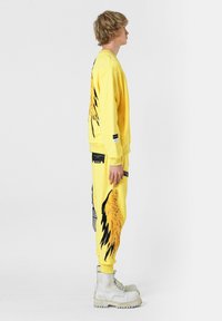 Sudadera amarilla y pantalones de chándal con un diseño de alas. Las características incluyen una textura suave, cuello redondo, puños elásticos y acentos en negro en contraste.