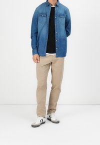 Jeansskjorta i blått med två bröstfickor, överlagd över en svart t-shirt. Beige byxor och vita sneakers med svarta ränder fulländar looken.