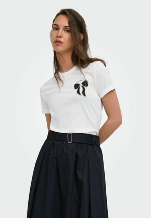 Donna che indossa una t-shirt bianca con una grafica di fiocco nero e testo "So Chic!", abbinata a una gonna plissettata nera a vita alta e cintura.