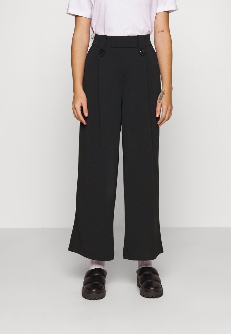 Svarta vida byxor med hög midja, med två framknappar, tillverkade av ett smidigt och lätt tyg. Kombinationen med mörka träskor och pastellfärgade strumpor.