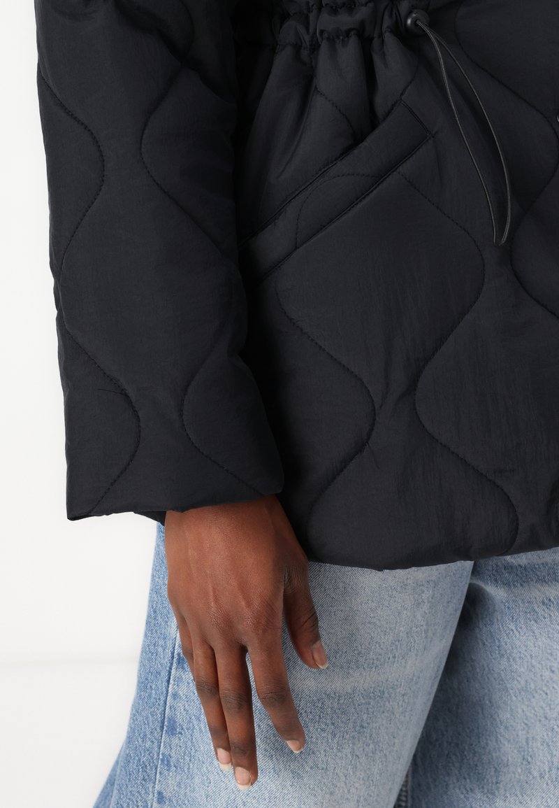 Schwarze gesteppte Jacke mit einem strukturierten Wellenmuster, elastischen Kordelzügen in der Taille und langen Ärmeln. Hand ruht auf Denimstoff.